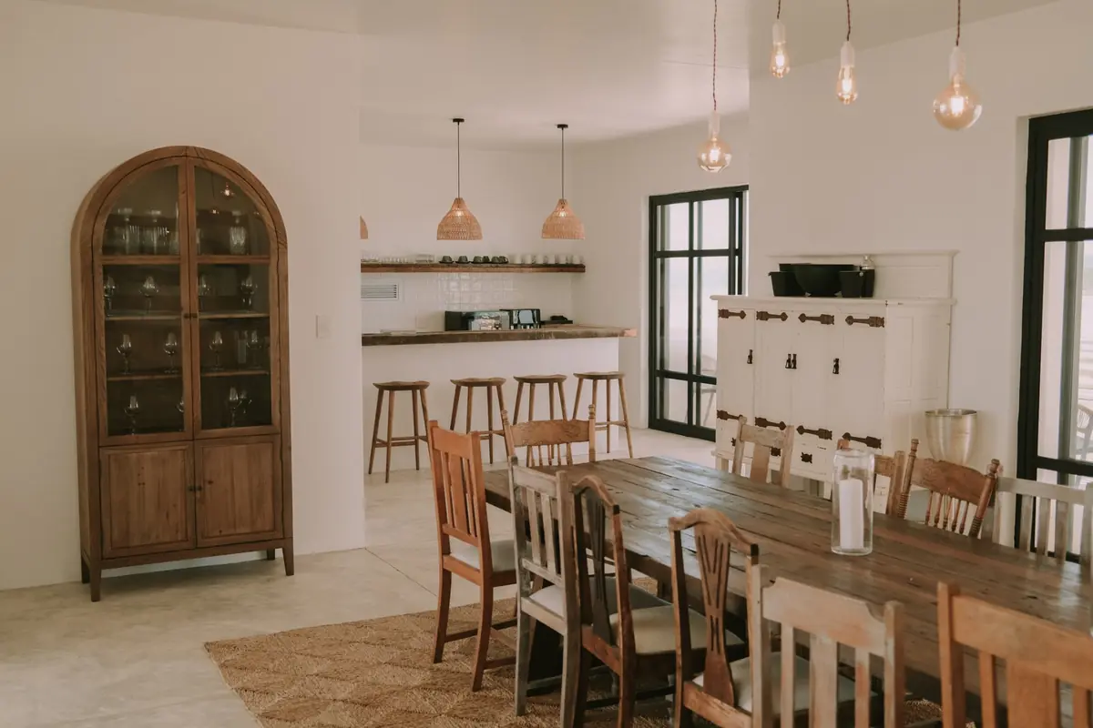 The long timber dining table with wishbone chairs, and the kitchen bar with rattan pendants, timber stools and an arched glass cabinet beyond