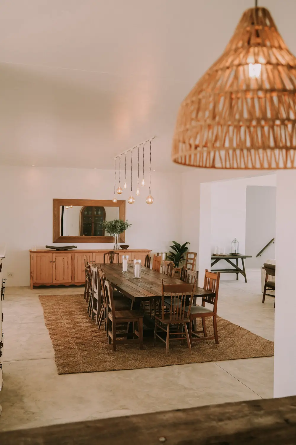 The long timber dining table on a seagrass rug, beneath a woven pendant light