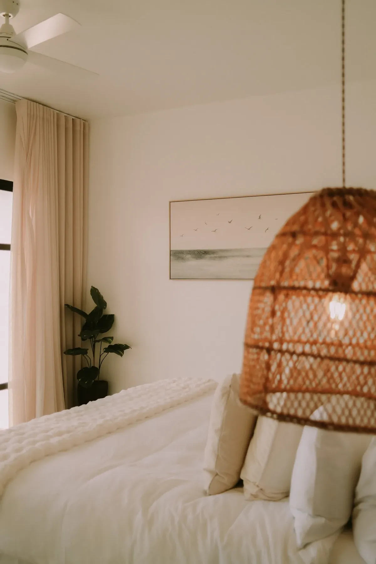 A bedroom with a rattan pendant light, a painting of seabirds and long curtains