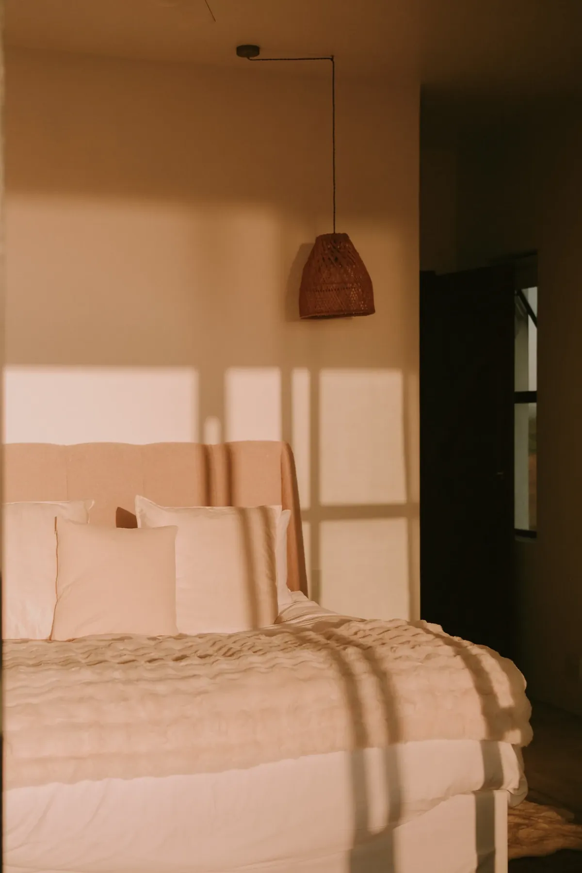 A bed beneath a rattan pendant light, with window shadows on the wall