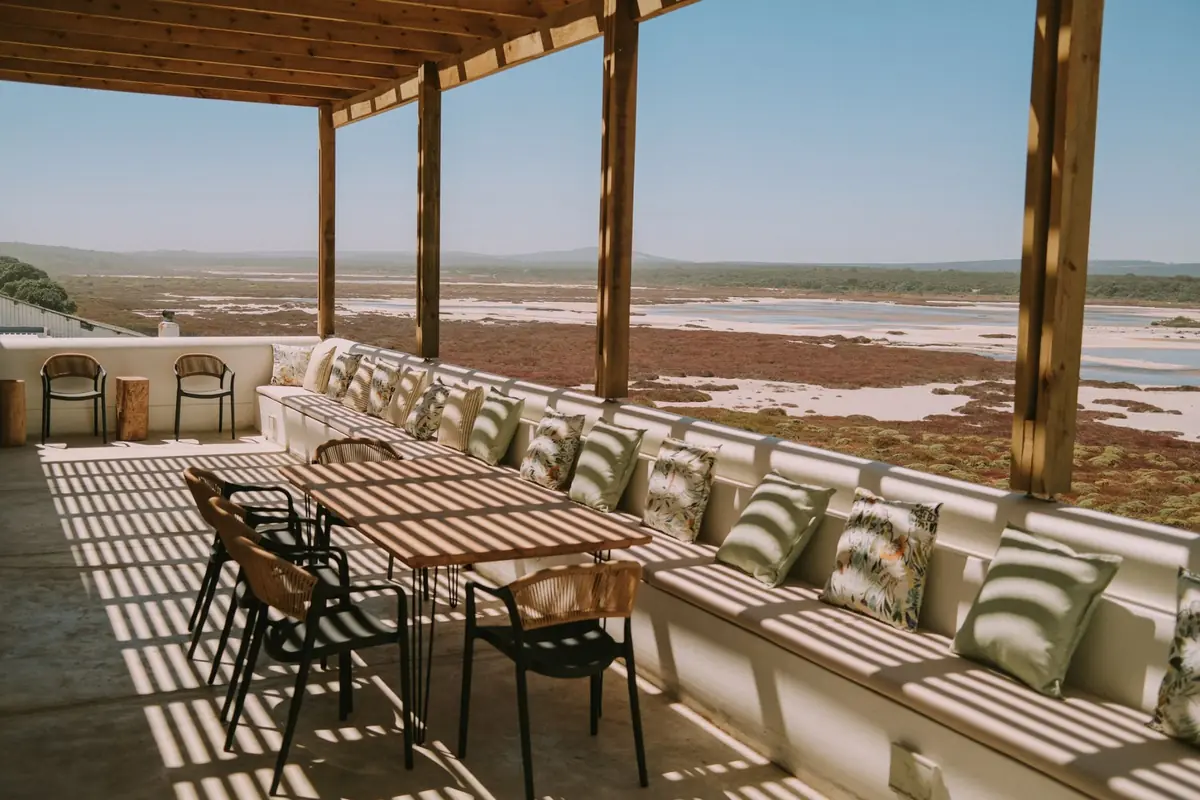 The covered terrace, with a long timber table and rattan chairs, and a built-in bench lined with cushions, looking over the salt pans