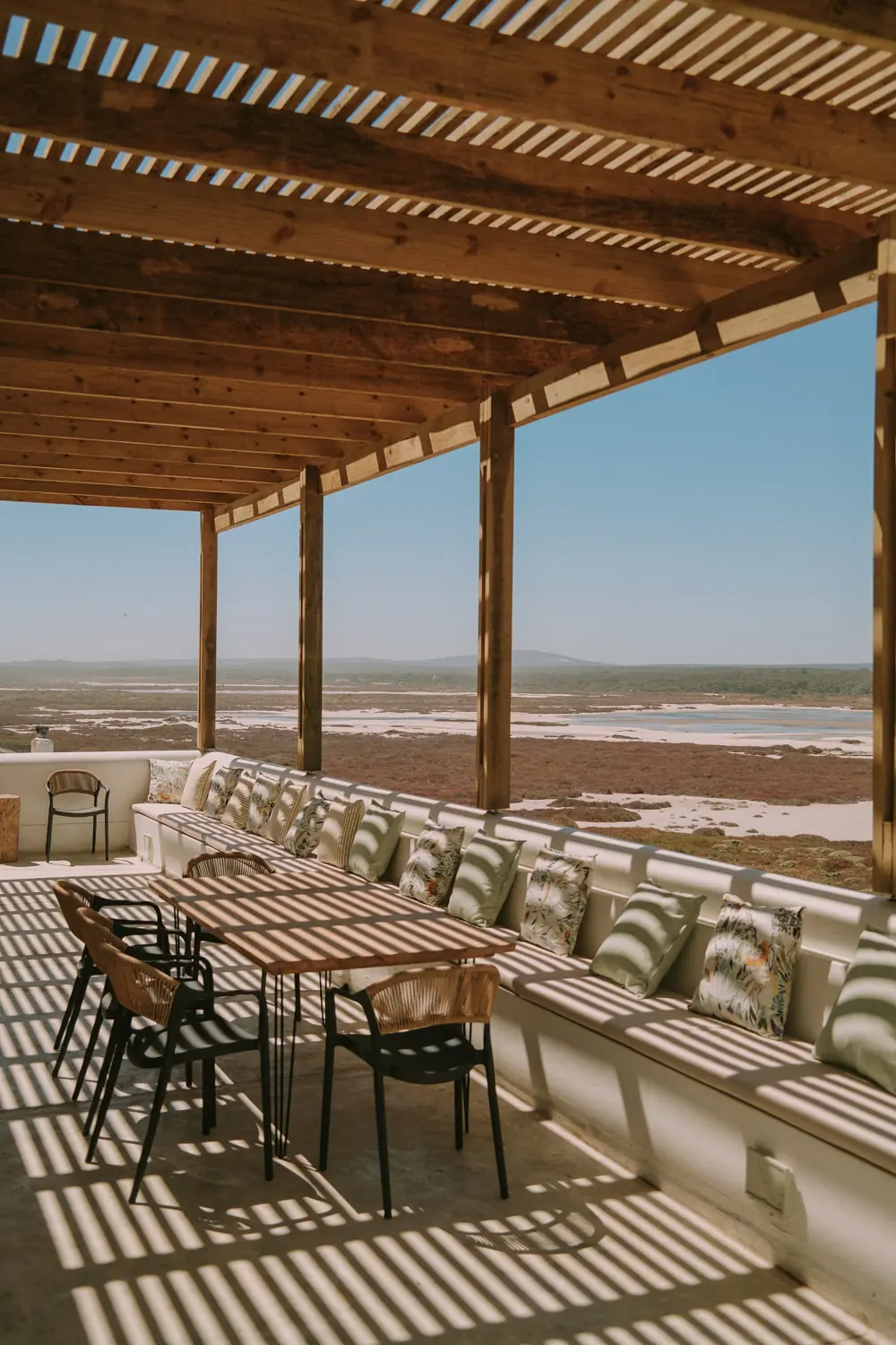 The covered terrace, with a long timber table, rattan chairs and a built-in bench in striped pergola shadows, looking out over the salt pans